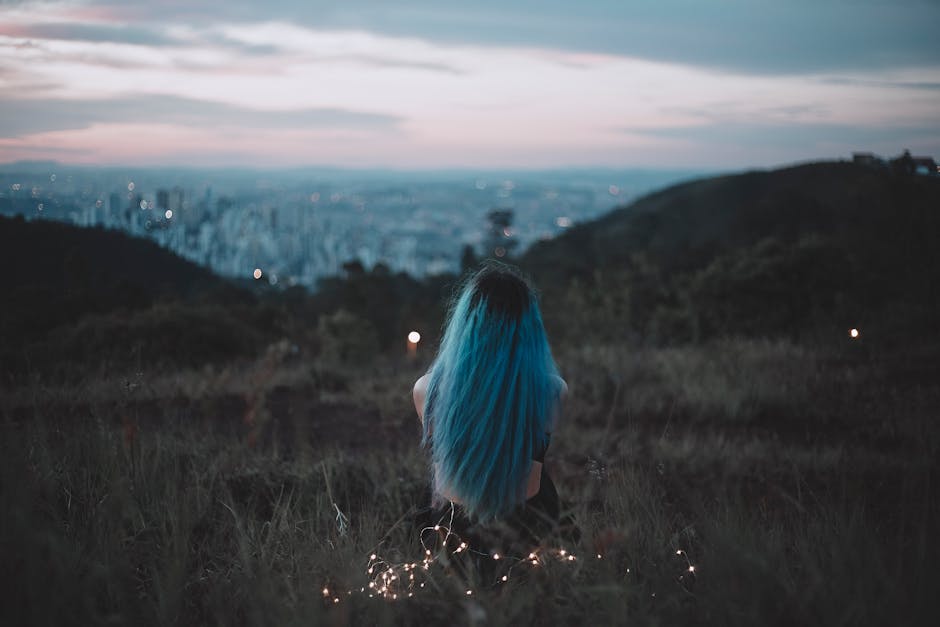 A woman with blue hair sits on a hillside, gazing at the city's twilight skyline adorned with string lights.